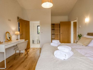A bedroom with a bed towels on it a desk chair lamp and vanity mirror and an open door showing a bathroom at Woodpecker Lodge in Butterleigh
