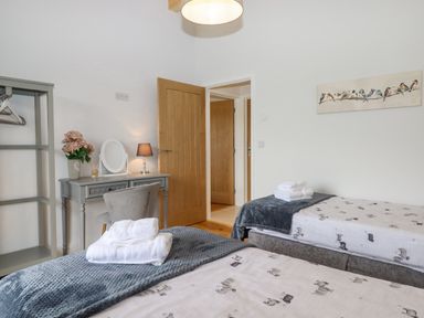 A bedroom with two single beds each with folded towels a grey blanket a grey dressing table with a chair a small lamp and flowers a wooden door and wall art of birds at Woodpecker Lodge in Butterleigh