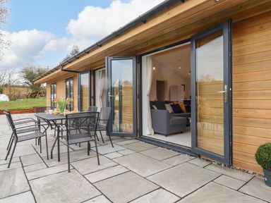An outdoor patio with a black metal dining table and chairs next to a wooden building with glass doors at Woodpecker Lodge in Butterleigh