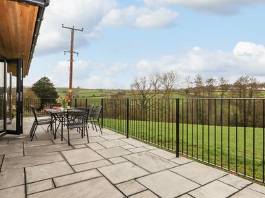 An outdoor patio with a dining table and chairs overlooking a grassy field at Woodpecker Lodge in Butterleigh
