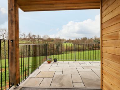A covered patio with stone tiles black metal railing and potted plants overlooking a green field at Woodpecker Lodge in Butterleigh