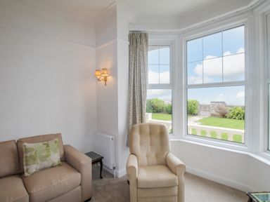 A sitting area with an armchair and a sofa near bay windows overlooking a garden at Weymouth Bay Apartment A in Weymouth