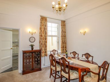 A dining room with a wooden dining table and six chairs set for a meal a cabinet with glassware a window with floral curtains and wall lights at Weymouth Bay Apartment A Weymouth