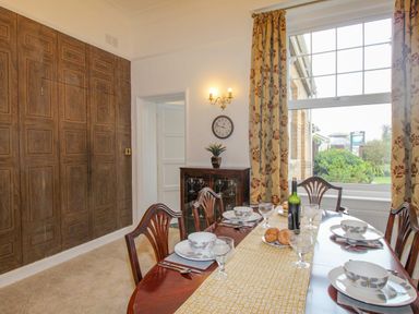 A dining room with a set table four chairs wood paneled wall side cabinet and window with floral curtains at Weymouth Bay Apartment A Weymouth