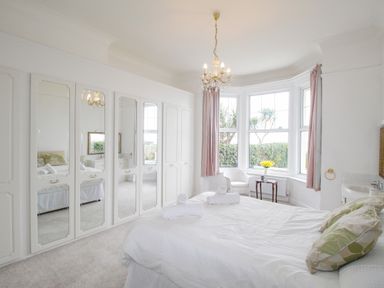 A bedroom with a bed white wardrobes with mirrors a chair a table with flowers and bay windows at Weymouth Bay Apartment A in Weymouth