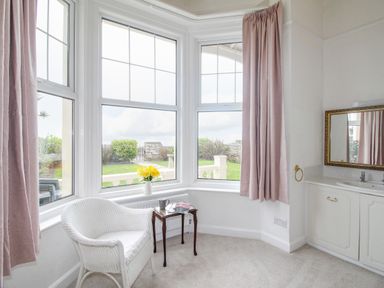 A room with large windows and pink curtains a white chair a small wooden table with a mug and flowers and a sink with a mirror at Weymouth Bay Apartment A in Weymouth