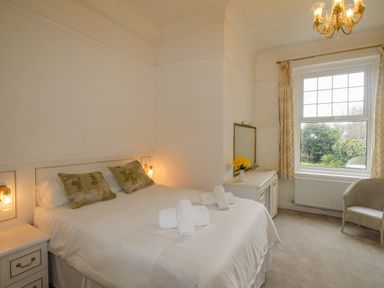 A bedroom with a double bed, bedside tables with lamps, a dresser with a mirror and flowers, a window with curtains, and a chair at Weymouth Bay Apartment A in Weymouth