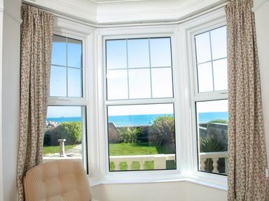 A window with three panes overlooking a garden and the sea with patterned curtains and a beige chair at Weymouth Bay Apartment A in Weymouth