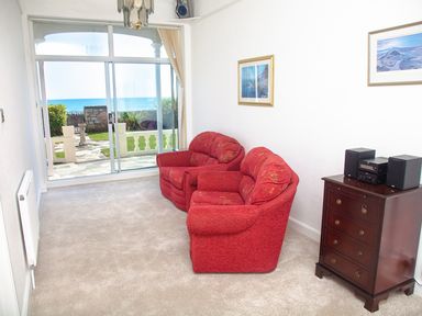A sitting room with two red armchairs a wooden chest of drawers with a stereo and paintings on the wall with sliding glass doors showing a garden and sea view at Weymouth Bay Apartment A in Weymouth
