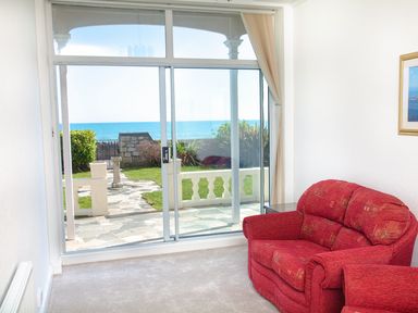 A sitting area with red armchairs facing a sliding glass door showing a garden and sea view at Weymouth Bay Apartment A Weymouth