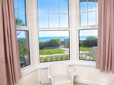 A white wicker chair by a bay window with pink curtains overlooking a garden and the sea at Weymouth Bay Apartment A in Weymouth