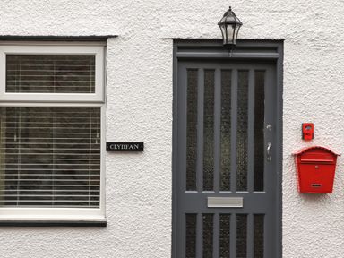 A grey front door with glass panels next to a window with blinds a red mailbox and a black house nameplate reading Clydfan on a white textured wall at Clydfan in Caernarfon