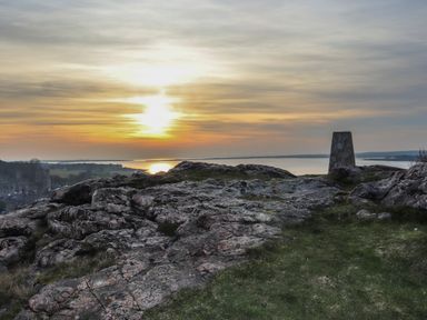 Rocky hill with a trig point overlooking a body of water at sunset with a castle in the distance at Clydfan in Caernarfon
