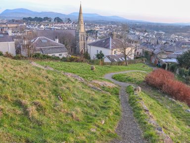 A winding pathway on a grassy hill overlooking houses and a church with mountains in the background at Clydfan in Caernarfon
