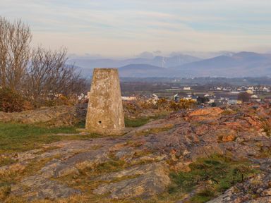 A rocky hill with a concrete triangulation pillar and a town with mountains in the background at Clydfan in Caernarfon