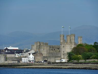A stone castle with flags near a body of water and buildings with mountains in the background at Clydfan in Caernarfon