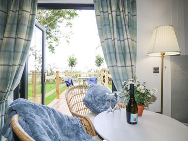 A small table with wine bottle and two wine glasses between two chairs with blue blankets next to a lamp and open door leading to an outdoor deck with chairs at High Rigg Shepherd's Sunset in Brampton Cumbria
