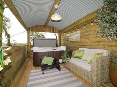 A covered outdoor space with a hot tub sofa with cushions a rug and a hanging light at High Rigg Shepherd's Sunset in Brampton Cumbria