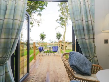 A deck with a round table and two chairs with blankets outside open glass doors with curtains at High Rigg Shepherd's Sunset in Brampton Cumbria