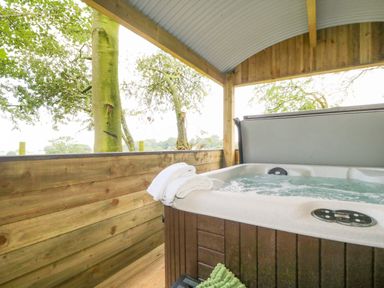 An outdoor hot tub with towels on the edge under a wooden shelter surrounded by trees at High Rigg Shepherd's Sunset in Brampton Cumbria