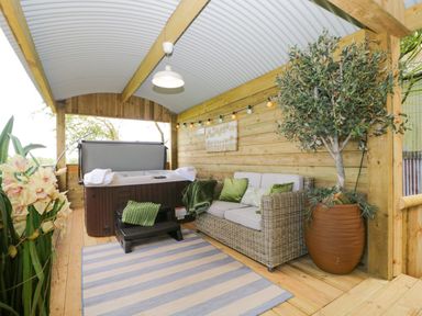 A covered outdoor seating area with a hot tub a sofa with green pillows and a large potted plant at High Rigg Shepherd's Sunset in Brampton Cumbria