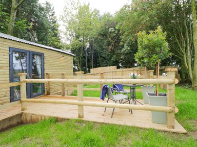 An outdoor wooden deck with a table and chairs next to a wooden building surrounded by trees at High Rigg Shepherd's Sunset in Brampton Cumbria