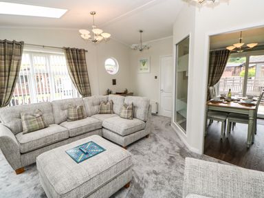 A living room with a grey sectional sofa, ottoman with a game board, plaid curtains, and a dining area with a table set at Badgers Retreat in Tunstall near Hipswell