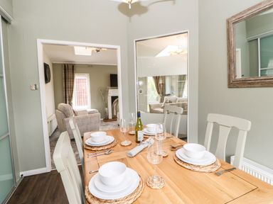 A dining room with a wooden table set with white dishes and glasses and chairs with a living room visible through an open doorway at Badgers Retreat in Tunstall near Hipswell