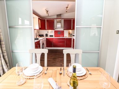 A dining table set for two with plates glassware and a bottle of wine looking into a kitchen with red cabinets at Badgers Retreat in Tunstall near Hipswell
