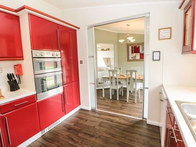 A kitchen with red cabinets and a double oven adjacent to a dining area with a wooden table and white chairs at Badgers Retreat in Tunstall near Hipswell