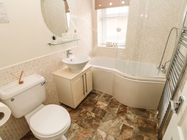 A bathroom with a toilet, sink on a cabinet, curved bathtub with glass screen, tiled walls, and a window with blinds at Badgers Retreat in Tunstall near Hipswell