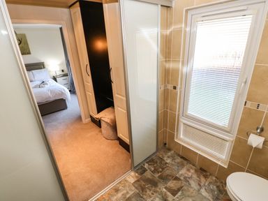 A bedroom with a bed and bedside table visible through a doorway from a bathroom with tiled floor and a window with blinds at Badgers Retreat in Tunstall near Hipswell