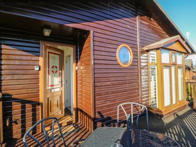 An outdoor patio with a glass table and metal chairs outside a wooden cabin with a door and circular window at Badgers Retreat in Tunstall near Hipswell