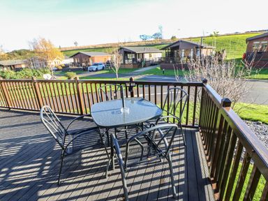 A wooden deck with a square glass table and four metal chairs overlooking a road and wooden cabins at Badgers Retreat in Tunstall near Hipswell