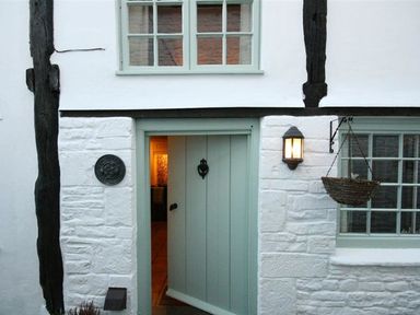 An exterior view of a white stone cottage with a half open light blue door and a lit lantern next to a window with a hanging basket at Butlers Cottage in Burford