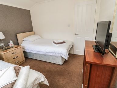 A bedroom with two single beds a wooden nightstand with a lamp a wooden dresser with a TV and a closed white door at Hillside View in Alnwick