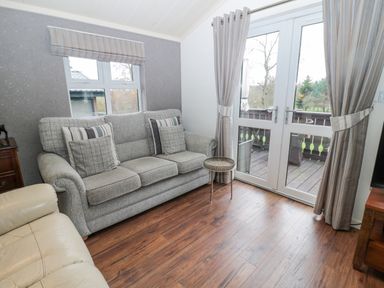 A living room with a gray sofa with cushions a small side table wooden flooring and glass double doors with curtains at Hillside View in Alnwick