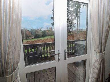 A view through glass double doors with curtains showing a wooden deck with a chair and a covered hot tub overlooking trees and grass at Hillside View in Alnwick