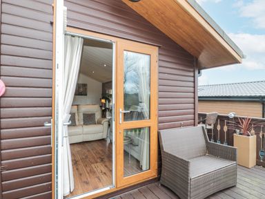 An outdoor patio area with a brown wicker loveseat a wooden deck and an open door leading to a living room with a beige sofa at Hillside View in Alnwick