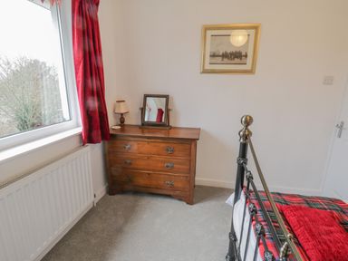 A bedroom corner with a wooden dresser holding a small mirror and lamp near a window with red curtains and a metal bed frame with red and plaid bedding at Sea Change in Newholm near Whitby