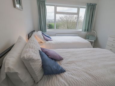 A bedroom with two single beds with white bedspreads and blue and purple pillows a window with curtains a white chair with cushions and a white chest of drawers at Sea Change in Newholm near Whitby