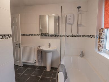 A bathroom with a pedestal sink, bathtub, shower, mirror cabinet, radiator, and a window with a seashell decoration at Sea Change in Newholm near Whitby