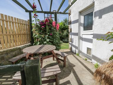 An outdoor patio area with a round wooden table and benches next to a white door and flowering plants at Sea Change in Newholm near Whitby