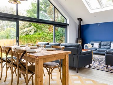 A dining area with a wooden table set with plates and glasses and a living area with dark sofas and a wood stove at The Garden House in Rock