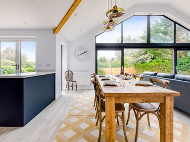 A dining area with a wooden table set with plates and glasses surrounded by chairs near large windows showing greenery at The Garden House in Rock