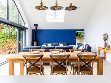 A dining area with a wooden table and six chairs in front of a living space with blue sofas and a blue accent wall at The Garden House in Rock