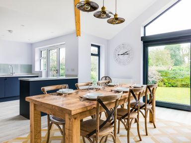 A dining area with a wooden table set with plates and glasses next to large windows and a kitchen at The Garden House in Rock