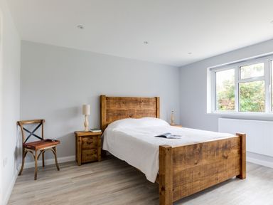 A bedroom with a wooden bed frame a white bedspread two bedside tables with lamps a wooden chair and a window at The Garden House in Rock