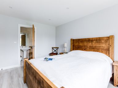 A bedroom with a wooden bed frame a white bedspread a wooden chair a wooden nightstand with a lamp and an open door showing a bathroom at The Garden House in Rock