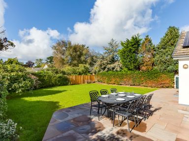 A patio with a dining table and six chairs set with plates and glasses next to a grassy garden with trees and shrubs at The Garden House in Rock